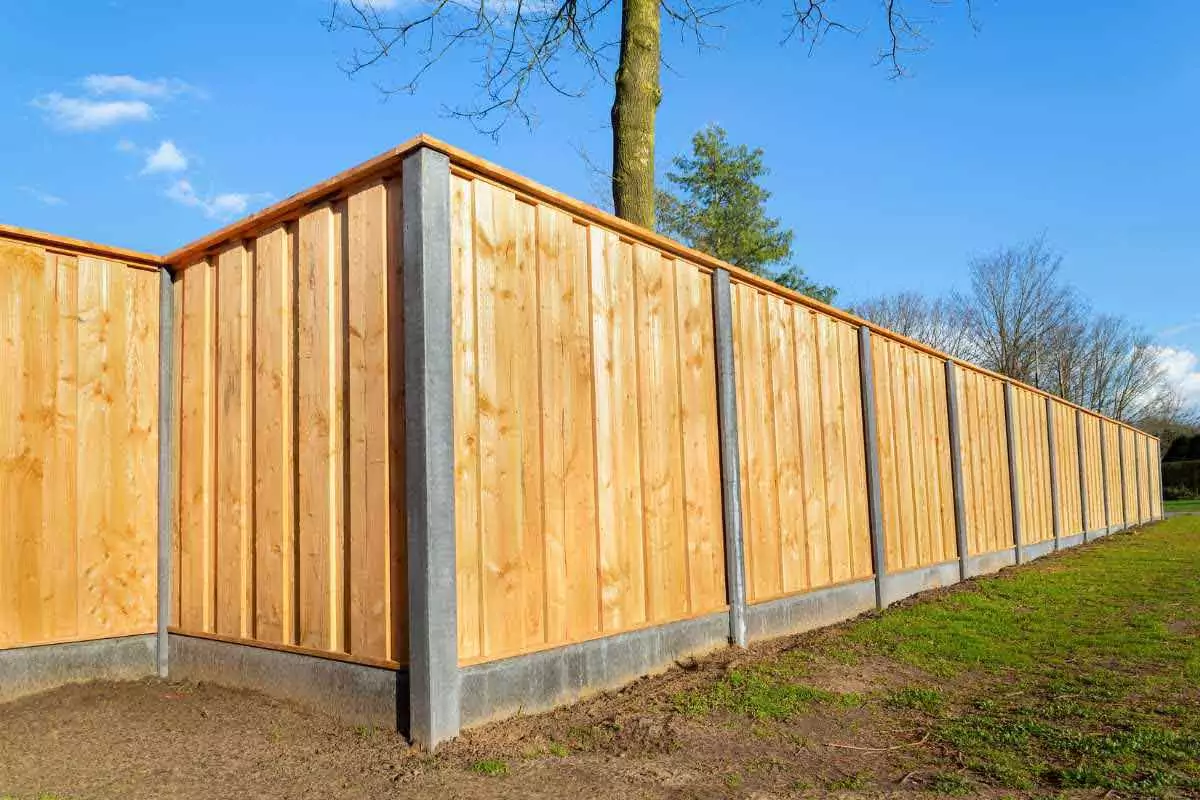 Wooden fence with concrete foundation and supports on a grass field, a tree behind it, under a clear blue sky.