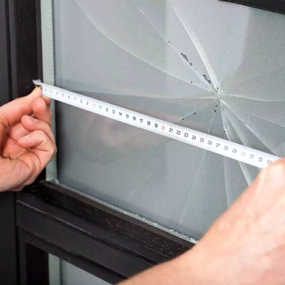 Man precisely measuring a broken glass pane on a window frame for replacement.