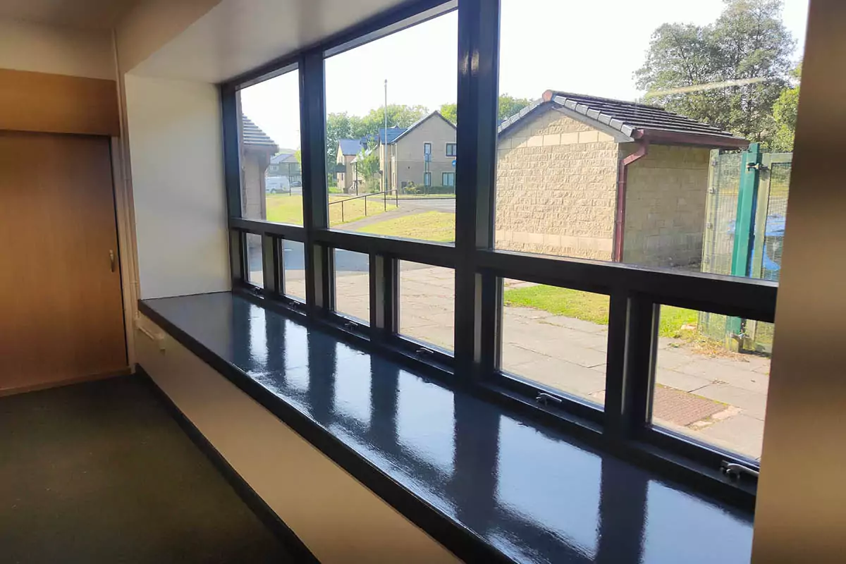 Interior view of a building with three large windows, each with two smaller windows below, black frames on a white wall, overlooking a sandstone building and modern houses.