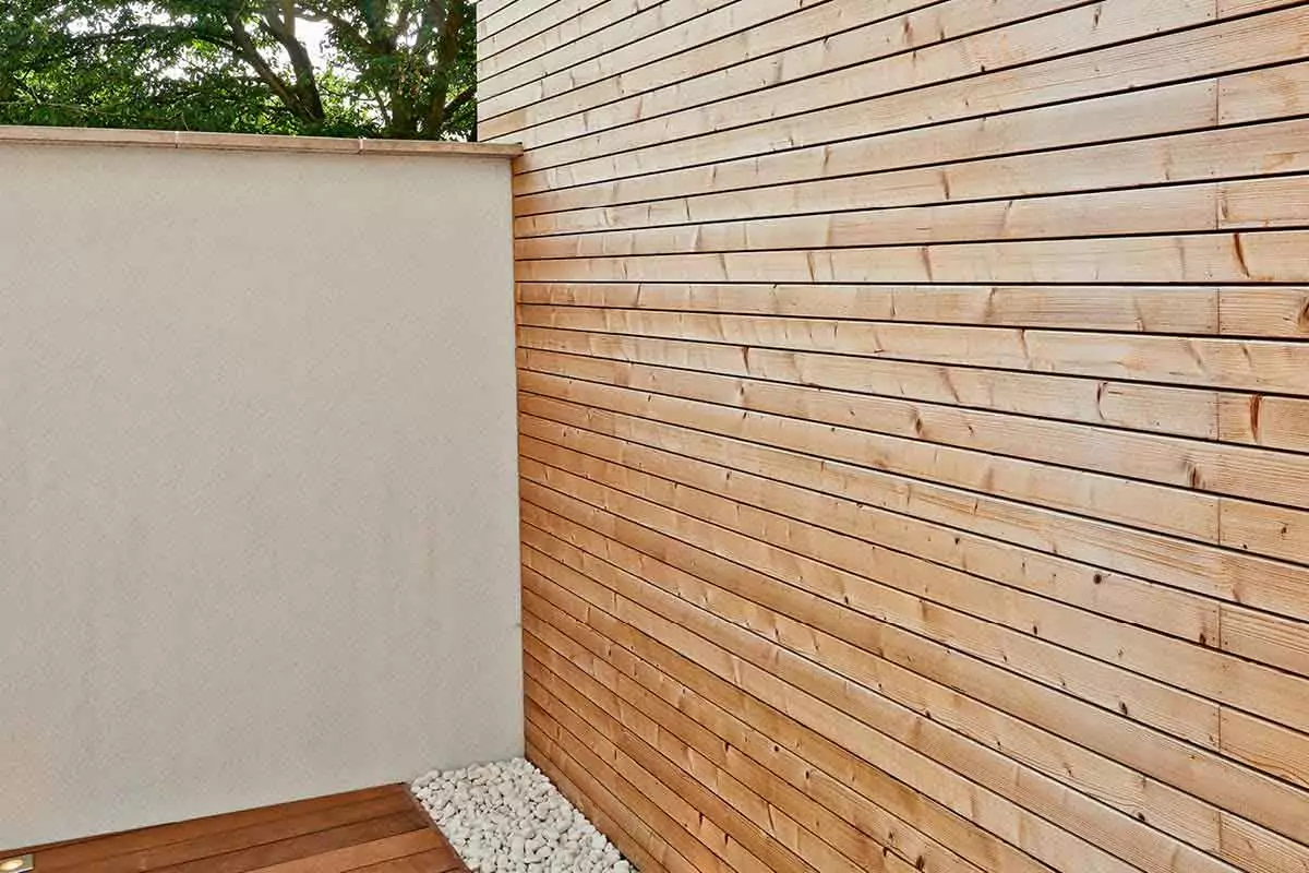 Outdoor scene with a white wall on the left, planked wood wall on the right, pebble stones, and wooden flooring.